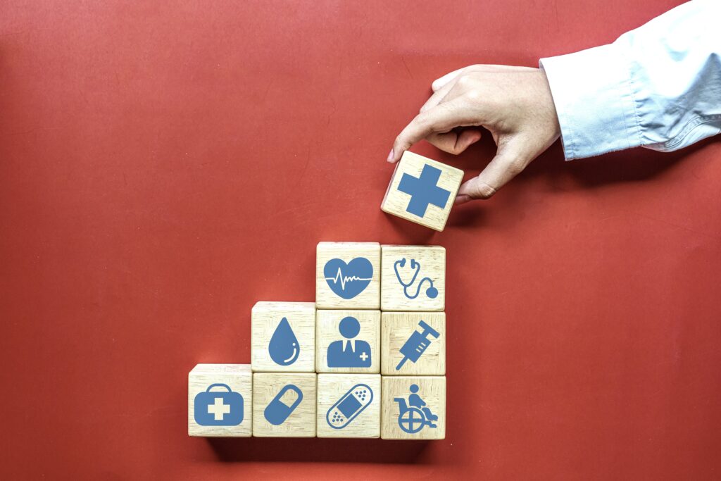 A hand places a wooden block with a medical cross on top of a stack of blocks. Other blocks display health symbols like a heart, stethoscope, syringe, and pill, arranged in a stair-step pattern against a red background.