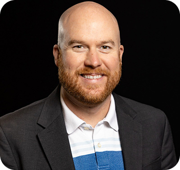 A smiling bald man with a red beard, wearing a dark blazer over a white collared shirt with blue and light blue stripes, posed against a plain black background.